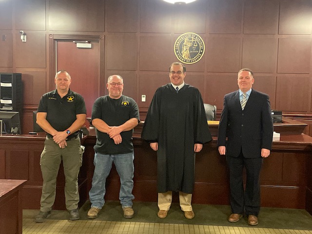 Constables, a judge, and other officials stand in front of a judge's desk.